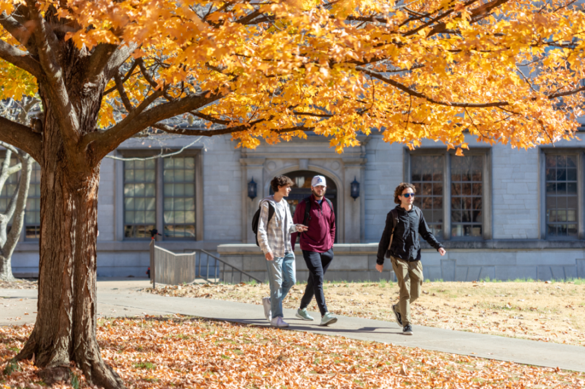 Three students walk in front of the Chemistry Building. It is fall and the leaves on the trees are orange and yellow.