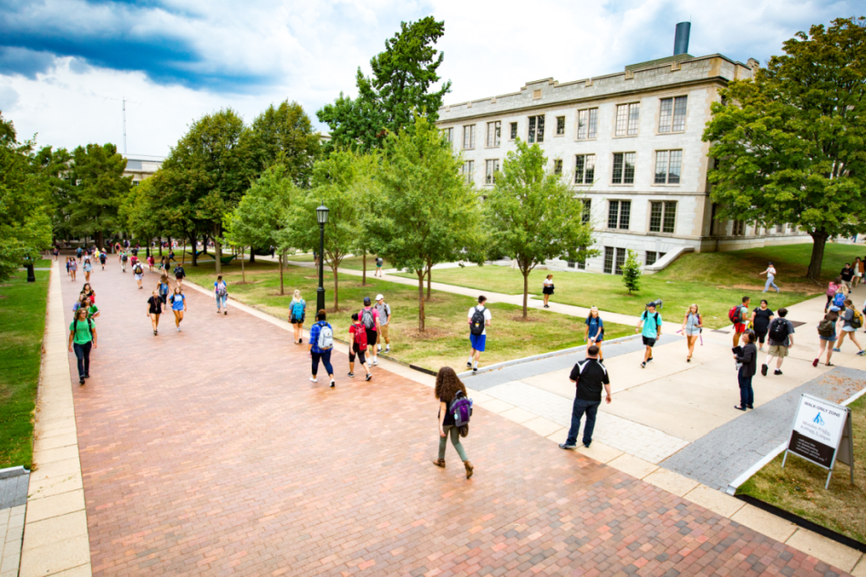 Students walk across campus. The Chemistry Building is in the background, and the trees on campus are bright green.